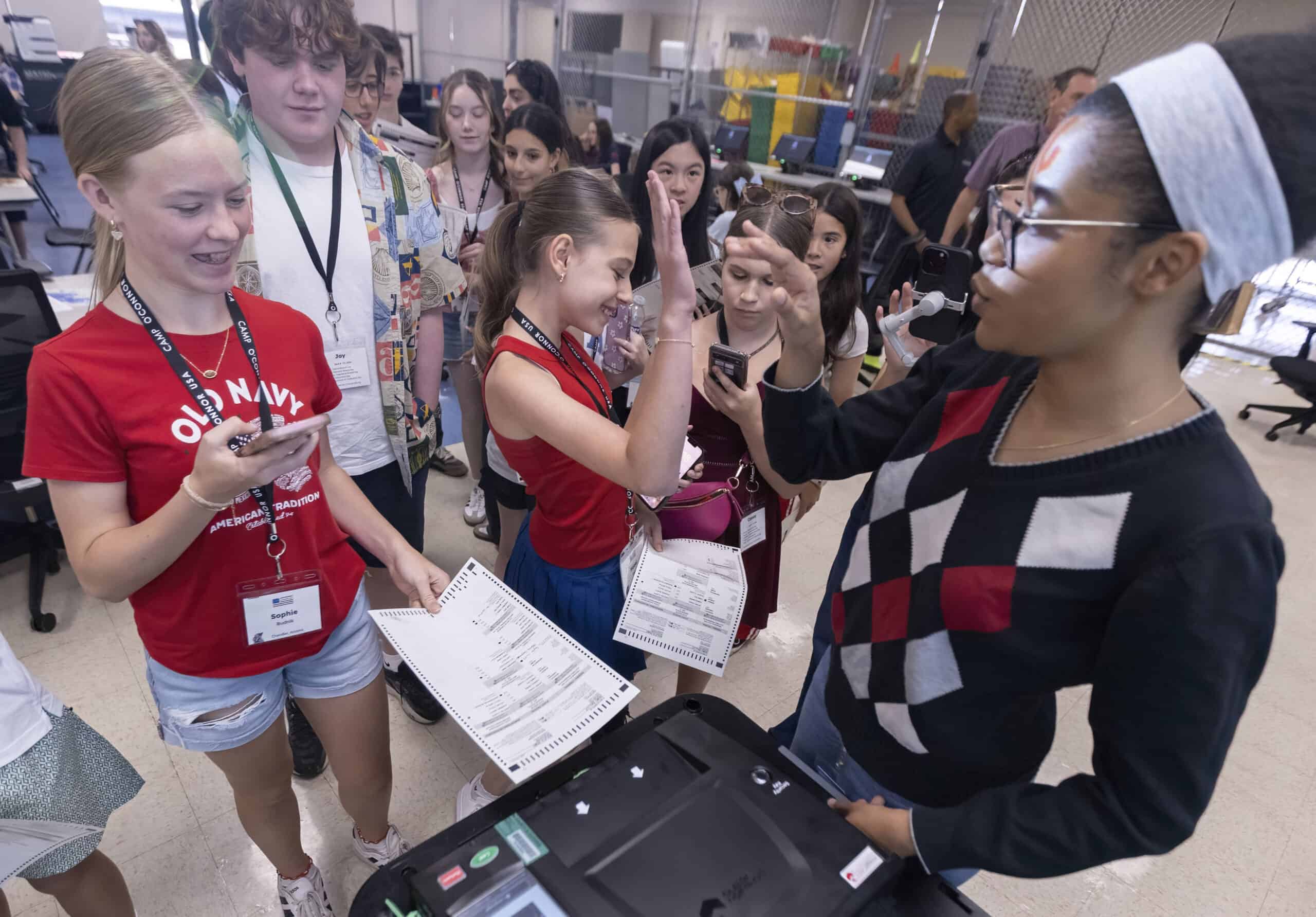 A group of students gather around a voting machine in a classroom. Two girls high-five while holding ballots, and others watch, smiling and using their phones. The vibrant scene captures the spirit of The Citizen in an engaging, educational setting.
