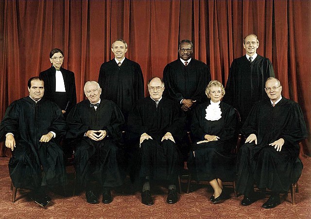 Nine people in black judicial robes pose in two rows before a red curtain; four are seated in front, five stand behind, all looking at the camera with the composed authority expected of those tasked with upholding The Citizen’s rights.