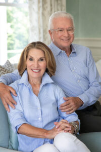 A smiling couple sits together indoors. Both are wearing blue shirts, with the woman seated and the man standing behind her, resting a hand on her shoulder. The background features light-colored furnishings and a window.