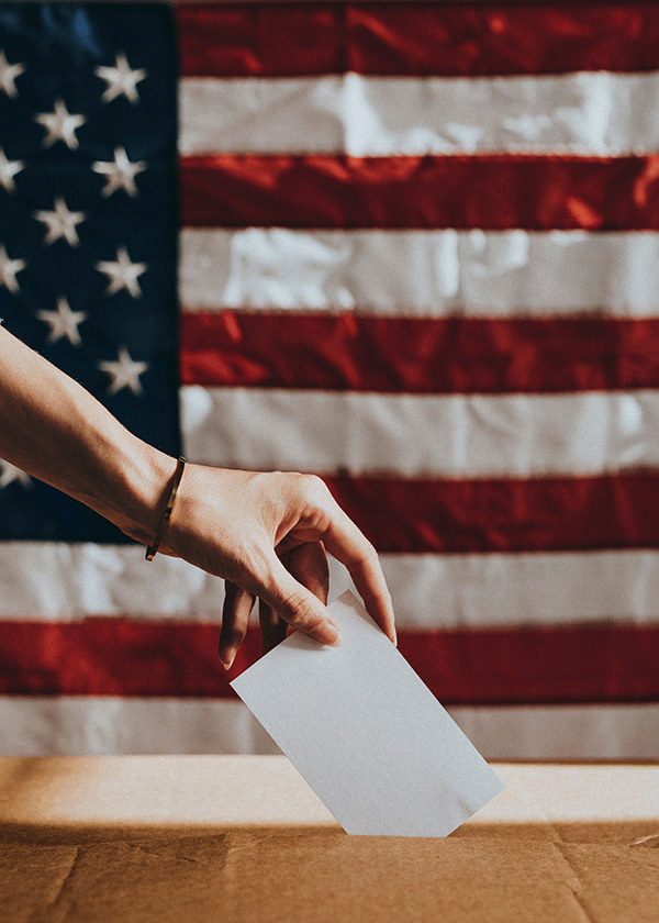 Voting A person’s hand places a white ballot into a box, with a large American flag in the background, highlighting the importance of Capital Connections in shaping democracy.