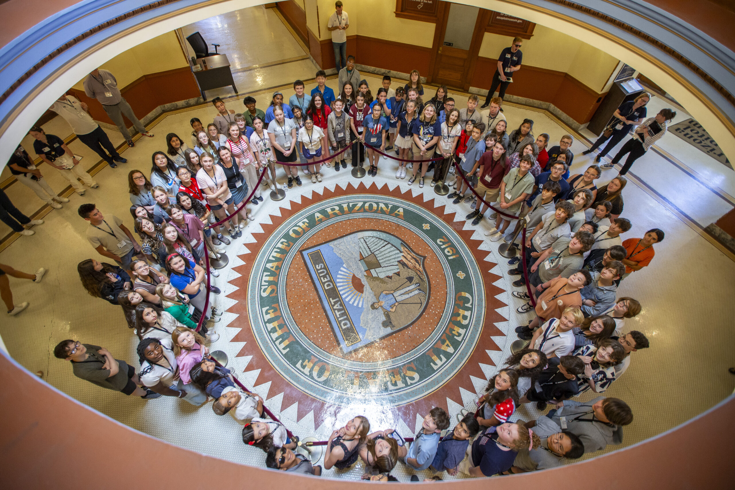 June 11, 2025 Camp SDO A large group of people, mostly children, stand in a circle around the Great Seal of the State of Arizona on the floor inside a government building, as each citizen looks up at the camera.