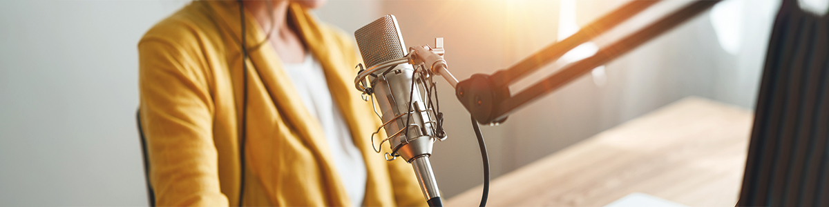 A person wearing a yellow jacket sits behind a microphone in a podcasting or broadcasting setup. The microphone is mounted on an adjustable stand, with sunlight streaming in from the side, illuminating the scene.