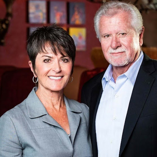 A woman with short dark hair and a gray blazer stands next to an older man with white hair and a beard, wearing a light blue shirt and dark jacket. They smile warmly in the warmly lit room, hinting at shared memories or perhaps stories of history over dinner, with artwork adorning the walls behind them.