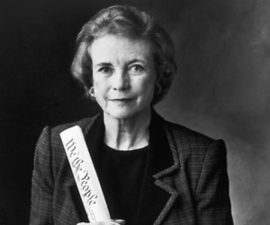 A black and white photo of a person in a checkered blazer holding a rolled-up paper with the words "We The People" visible. The background is dark and unadorned.