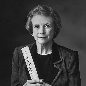 We the People_Web335x336 An elderly woman with short, wavy hair, wearing a checkered blazer, holds a rolled document. The black and white image has a subtle, serious tone. The document has partially visible text.