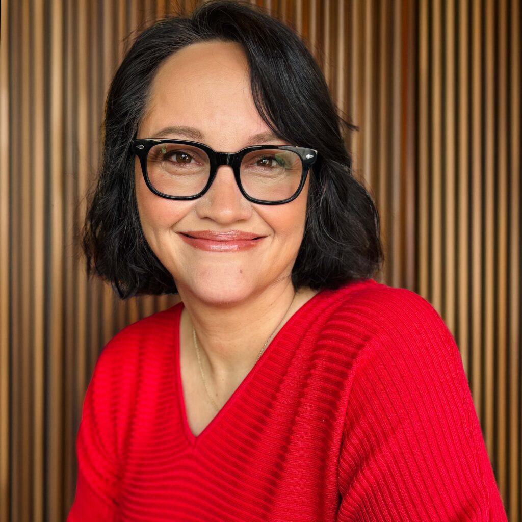 A woman with short dark hair and glasses smiles at the camera, wearing a bright red sweater, with a background of vertical wooden slats.