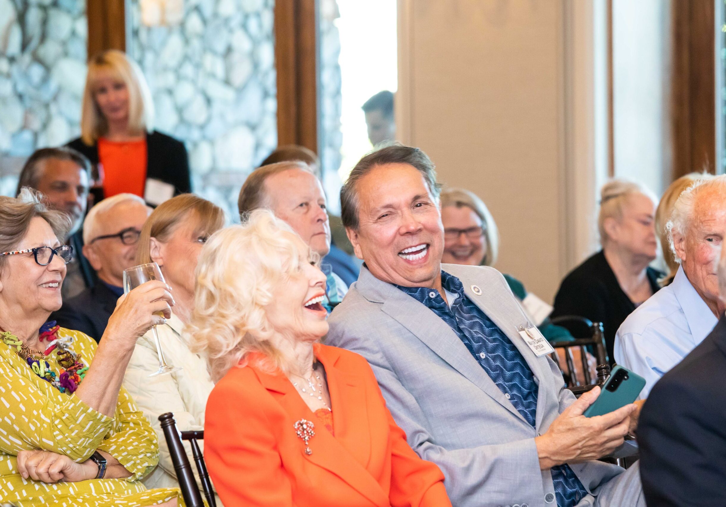 A group of elderly people sitting and smiling at an indoor event. A man in a light gray suit is holding a smartphone, and a woman in an orange blazer is laughing beside him. Others are seated around them, some holding glasses.