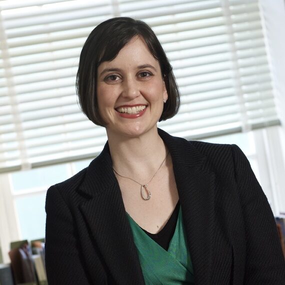 A person with short dark hair is smiling, wearing a black blazer over a green top, with a necklace. They are standing in front of a window with blinds partially open, letting in light.