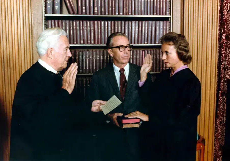 A woman, like Sandra Day O'Connor, stands with her right hand raised and left hand on a book, being sworn in by a man in a judge’s robe. Another man stands between them holding a card, with law books filling the bookshelf behind them.