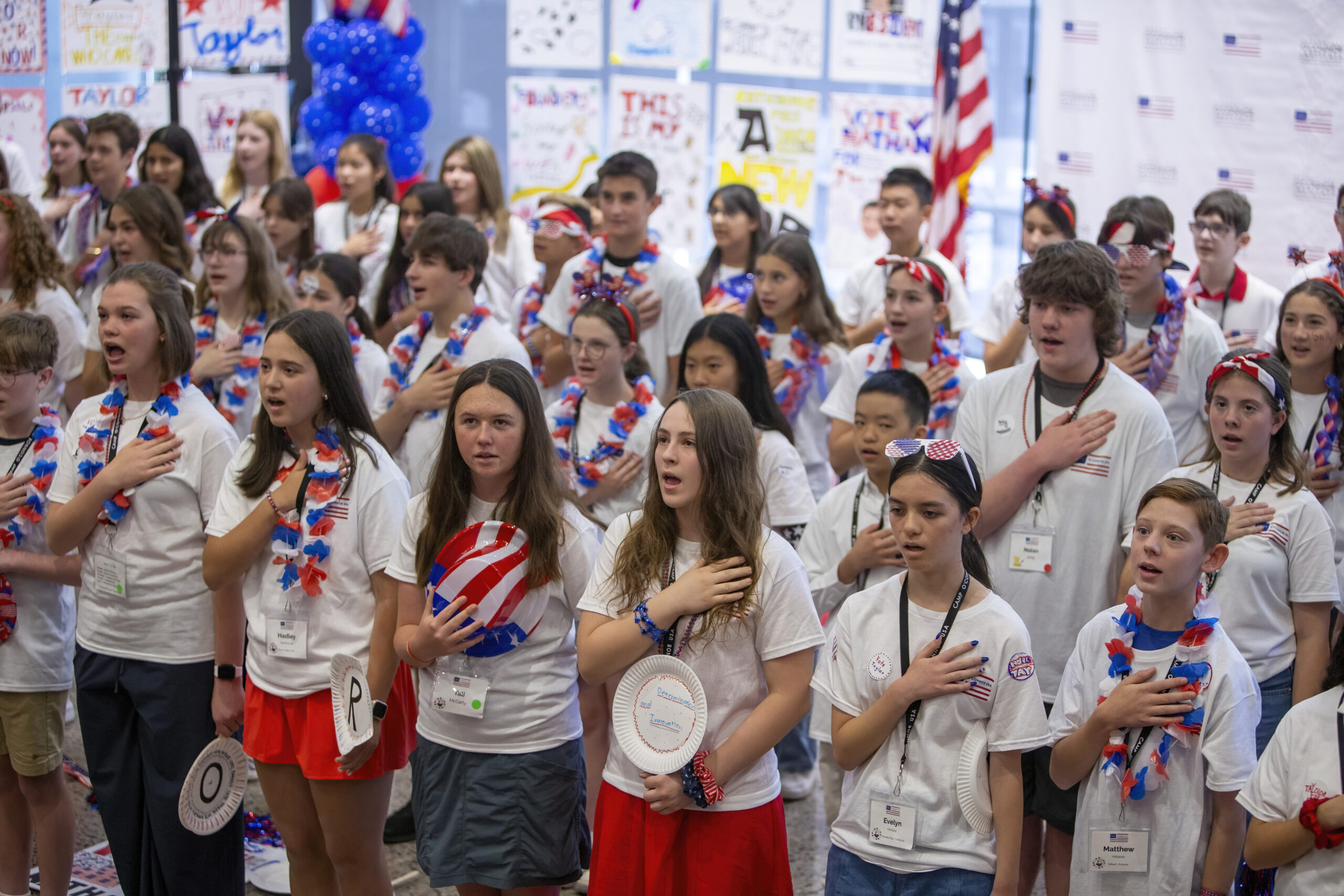 June 13, 2025 Camp SDO A large group of young citizens wearing red, white, and blue clothing and accessories stand with their right hands over their hearts, appearing to recite the Pledge of Allegiance at a patriotic event. Patriotic posters and an American flag are visible.