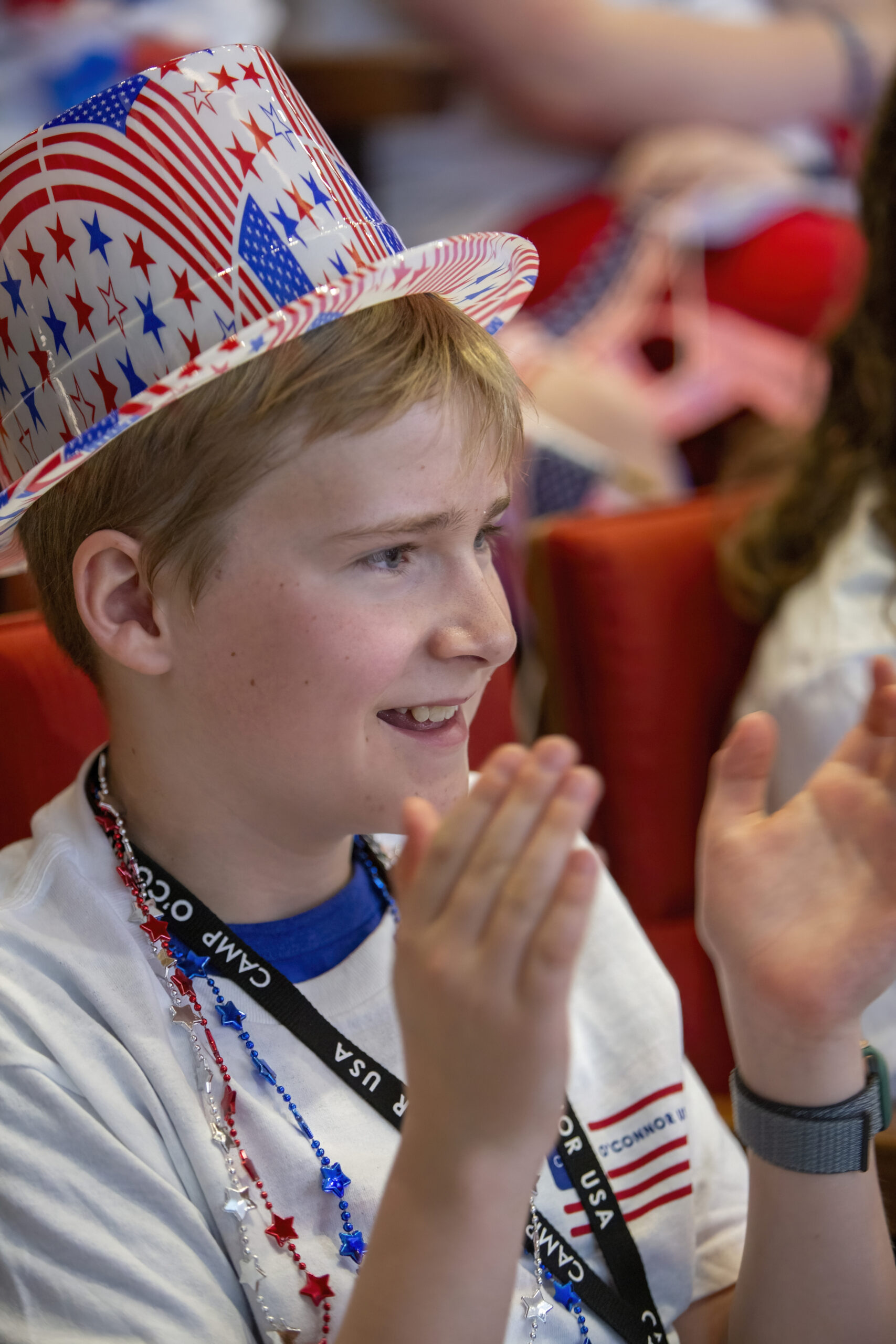 June 13, 2025 Camp SDO A smiling child wearing an America 250 patriotic hat decorated with stars and stripes claps hands at an event. The child wears a white shirt, a lanyard, and festive red, white, and blue beads.