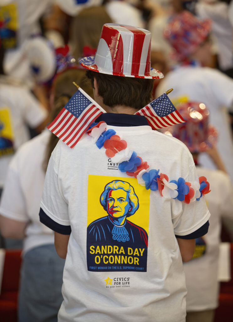 June 13, 2025 Camp SDO A citizen wearing a patriotic hat and a shirt with an image of Sandra Day O’Connor stands in a crowd. The shirt features red, white, and blue colors, with two small American flags tucked into the collar.