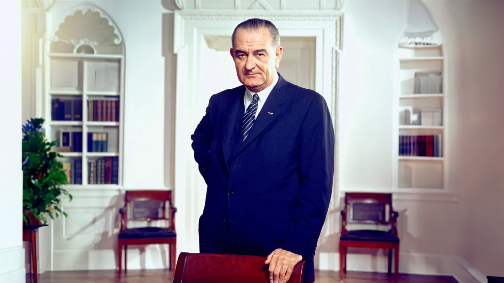 An older man in a suit stands confidently in an elegant room with white walls and ornate details, reminiscent of a grand history. Wooden chairs form the backdrop as he rests one hand on a chair, exuding poise as if poised for an important dinner. The room is softly lit.