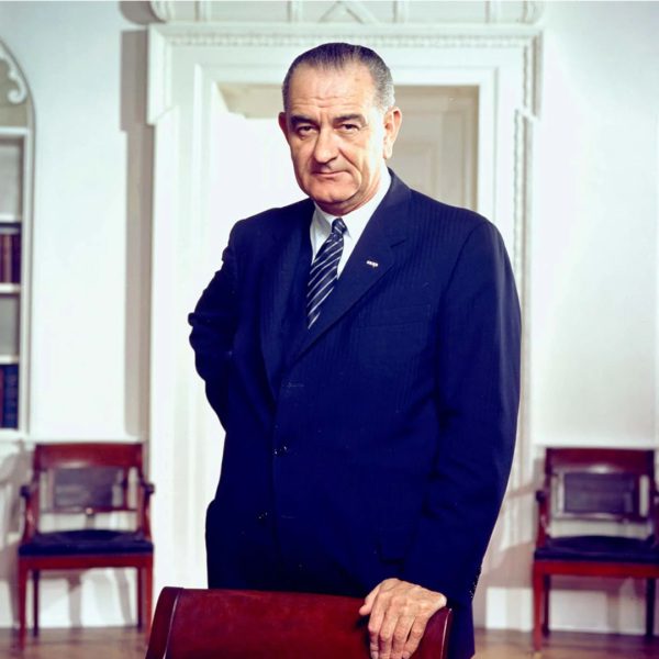An older man in a suit stands confidently in an elegant room with white walls and ornate details, reminiscent of a grand history. Wooden chairs form the backdrop as he rests one hand on a chair, exuding poise as if poised for an important dinner. The room is softly lit.