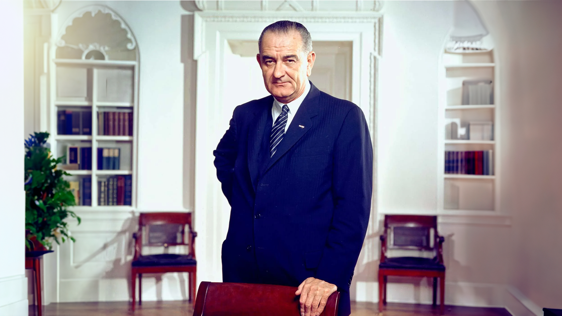 full room An older man in a suit stands confidently in an elegant room with white walls and ornate details, reminiscent of a grand history. Wooden chairs form the backdrop as he rests one hand on a chair, exuding poise as if poised for an important dinner. The room is softly lit.