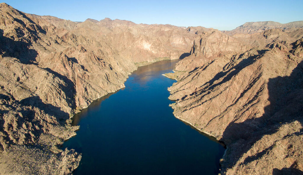 Aerial view of the Colorado River flowing between rocky, tan-colored canyon walls under a clear blue sky. The river's dark blue surface contrasts with the rugged, sunlit desert terrain surrounding it.