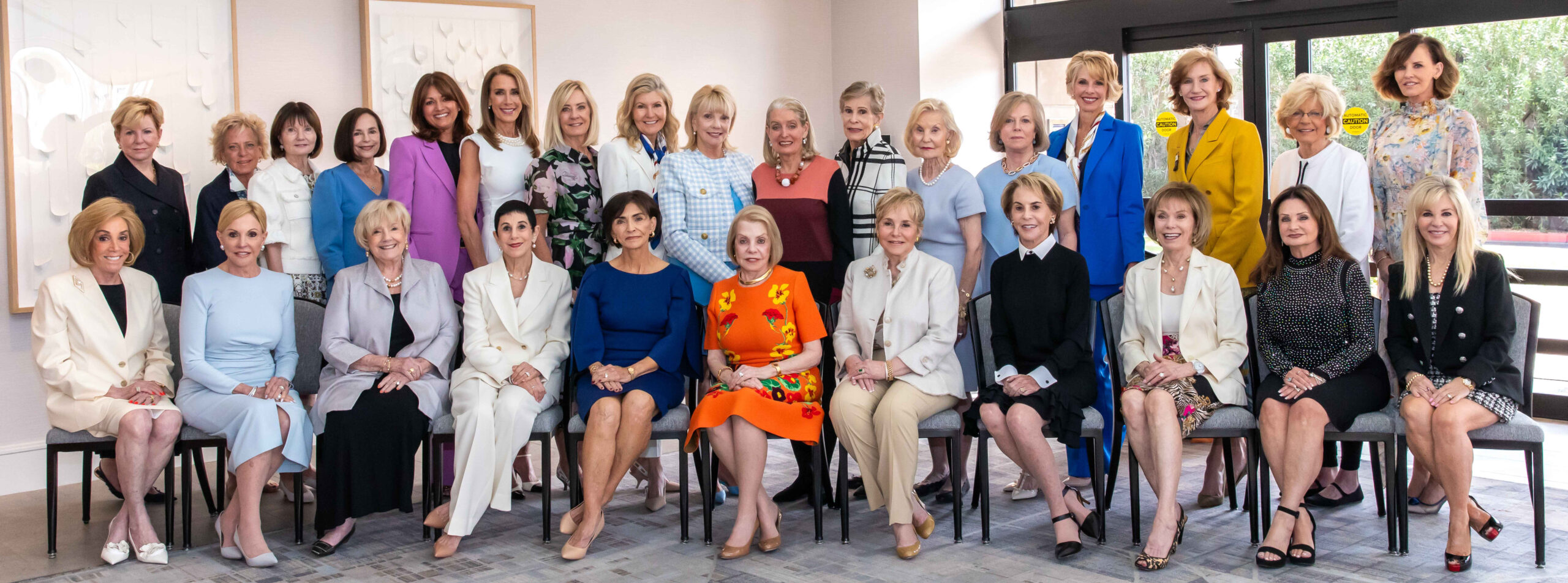 A group of women in leadership poses together indoors at a legacy luncheon. They are dressed in a variety of colorful and formal outfits, sitting and standing in several rows. The setting appears to be a well-lit room with framed art on the walls.