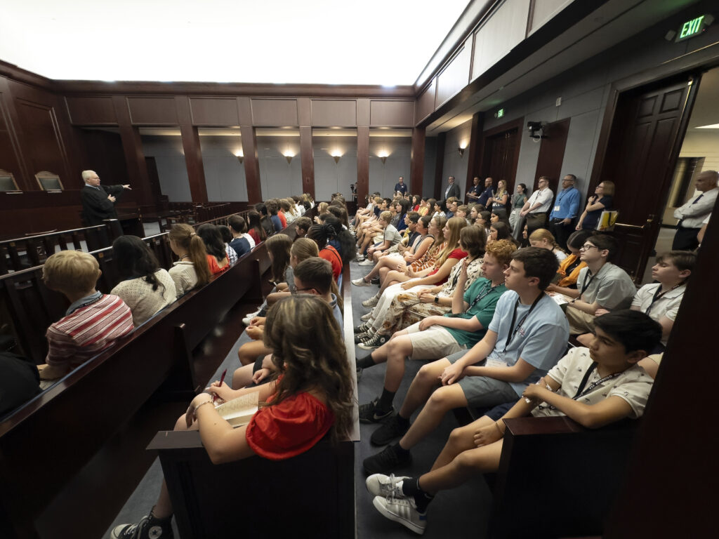 A large group of students sit attentively in wooden benches inside a courtroom, experiencing Our Impact as they listen to a speaker at the front. Adults stand along the walls, observing the well-lit session in the high-ceilinged room.