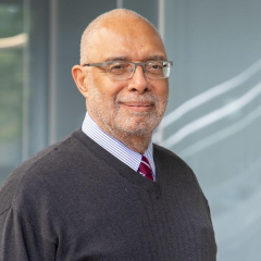 An older man with glasses and a gray beard smiles slightly. He is wearing a dark sweater over a striped shirt and tie. The background is a blurred, gray interior with glass windows.