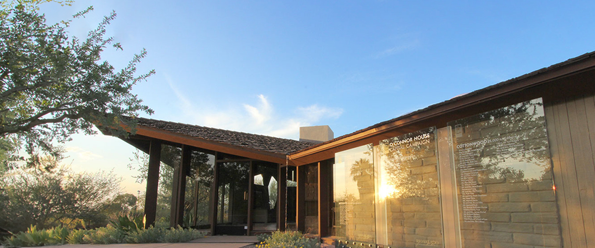 A modern architectural house with large glass windows reflecting sunlight, surrounded by trees. The sky is clear with a few clouds, creating a serene setting.