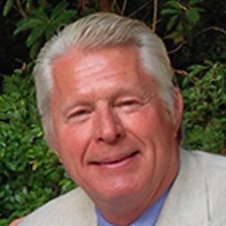 An older man with light hair and a smile wears a light-colored blazer and blue shirt. He is outdoors with green foliage in the background.