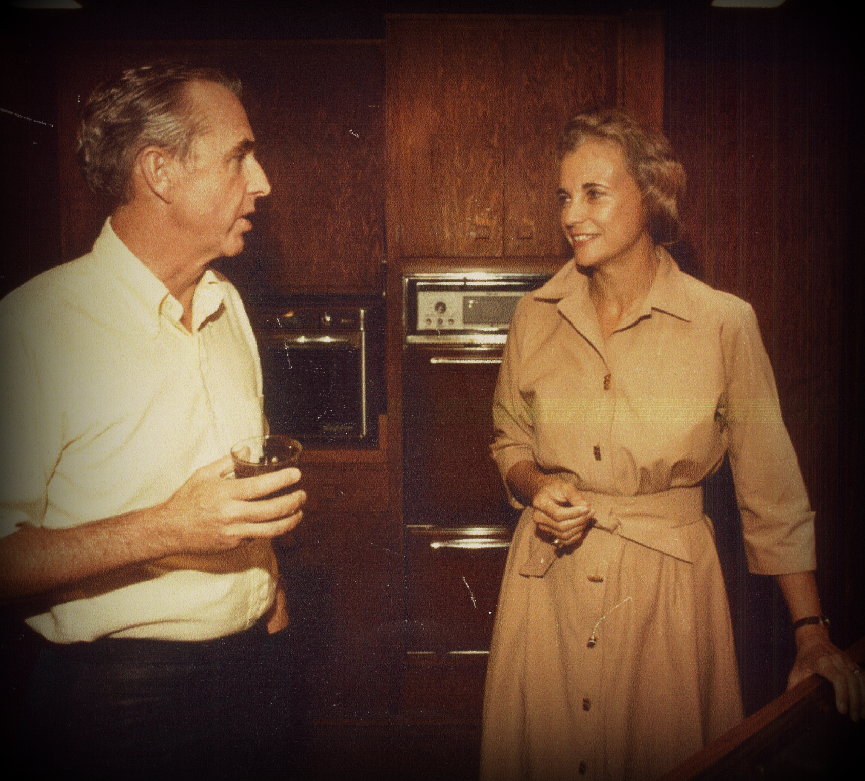 A man and woman stand in a kitchen having a conversation. The man holds a drink in his hand, while the woman wears a light-colored dress. The kitchen features wooden cabinets and an oven in the background. The photo has a vintage appearance.