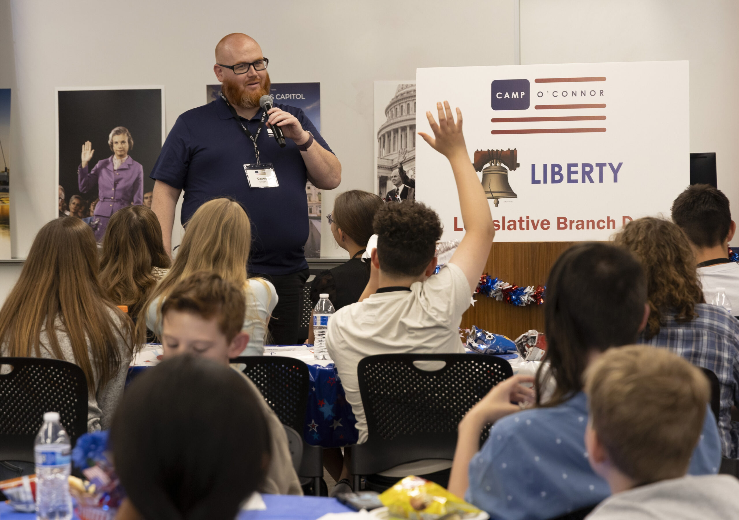 June 11, 2025 Camp SDO A man speaks into a microphone in front of a group of seated children, one of whom—an eager citizen in the making—has their hand raised. A sign behind him reads “Camp O’Connor Liberty Legislative Branch.”.
