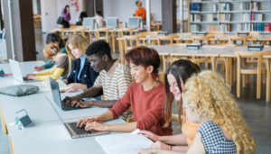 A diverse group of students is seated at a long table in a library, using laptops and reading. Shelves of books are visible in the background. The atmosphere is focused and collaborative.